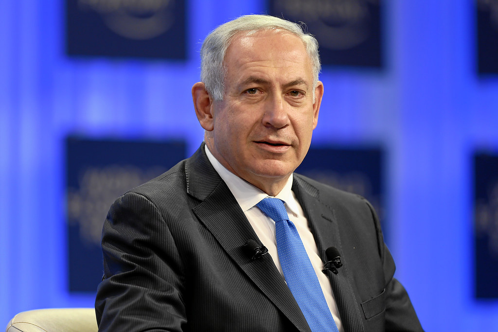 Benjamin Netanyahu in a dark suit and blue tie, seated against a blue background